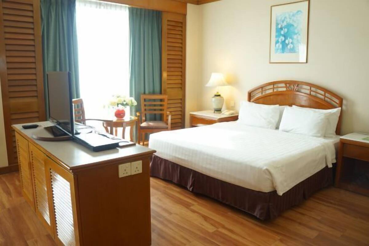 A hotel room with a wooden desk, a bed with white linens, a chair, a lamp, and a painting on the wall.