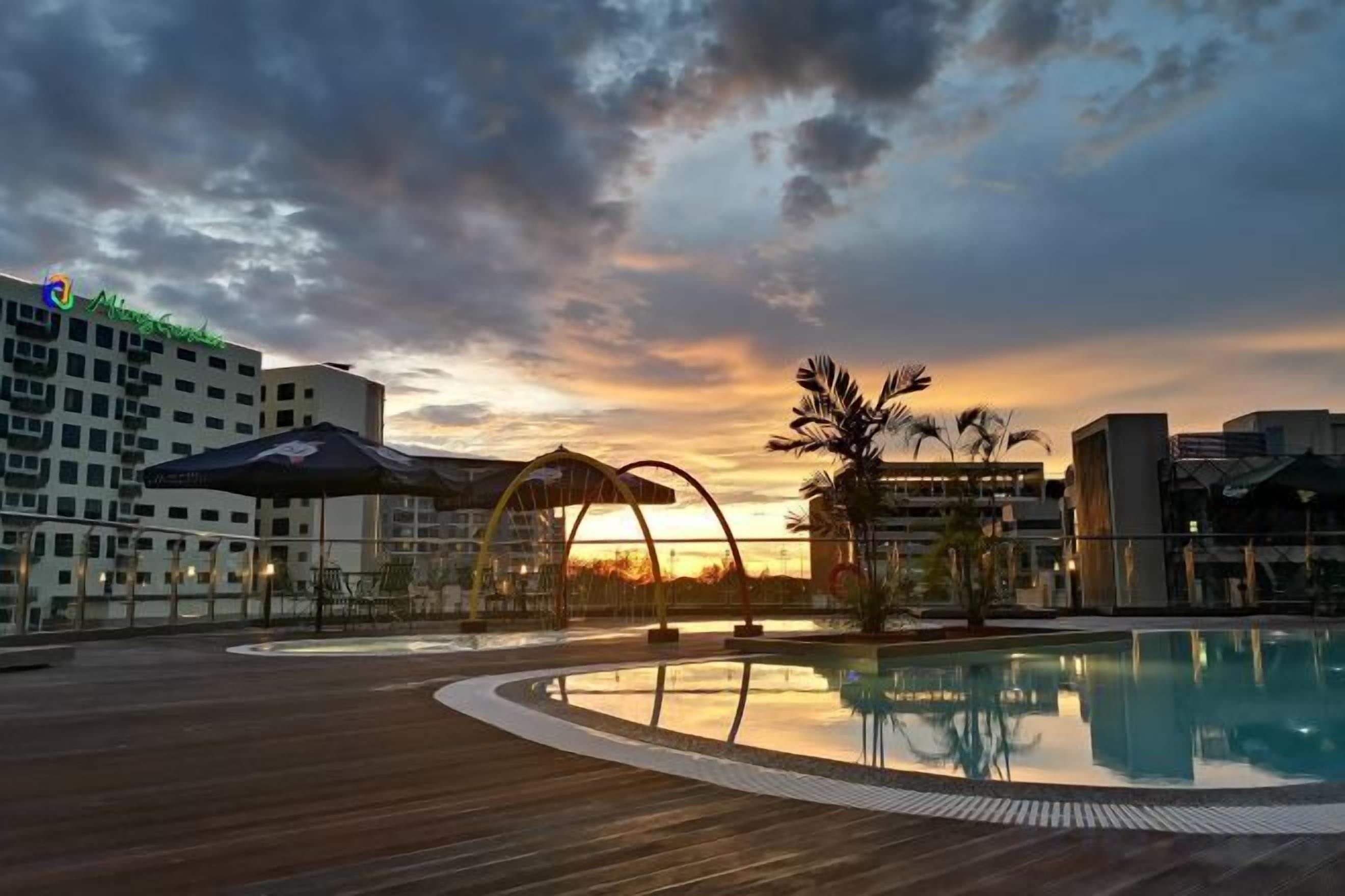 A rooftop pool with a wooden deck, surrounded by buildings and palm trees, reflecting the sunset.