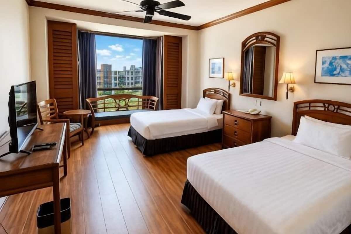 A hotel room with two beds, a desk, a chair, a TV, a ceiling fan, and a window with a view of buildings.