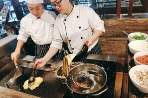 Deux chefs préparent des plats sur un grill, avec divers ingrédients et ustensiles de cuisine à portée de main.