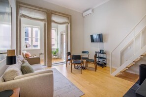 A modern living room with a sofa, a dining table with chairs, a TV mounted on the wall, and a staircase. - ALMADA 151 (Porto)