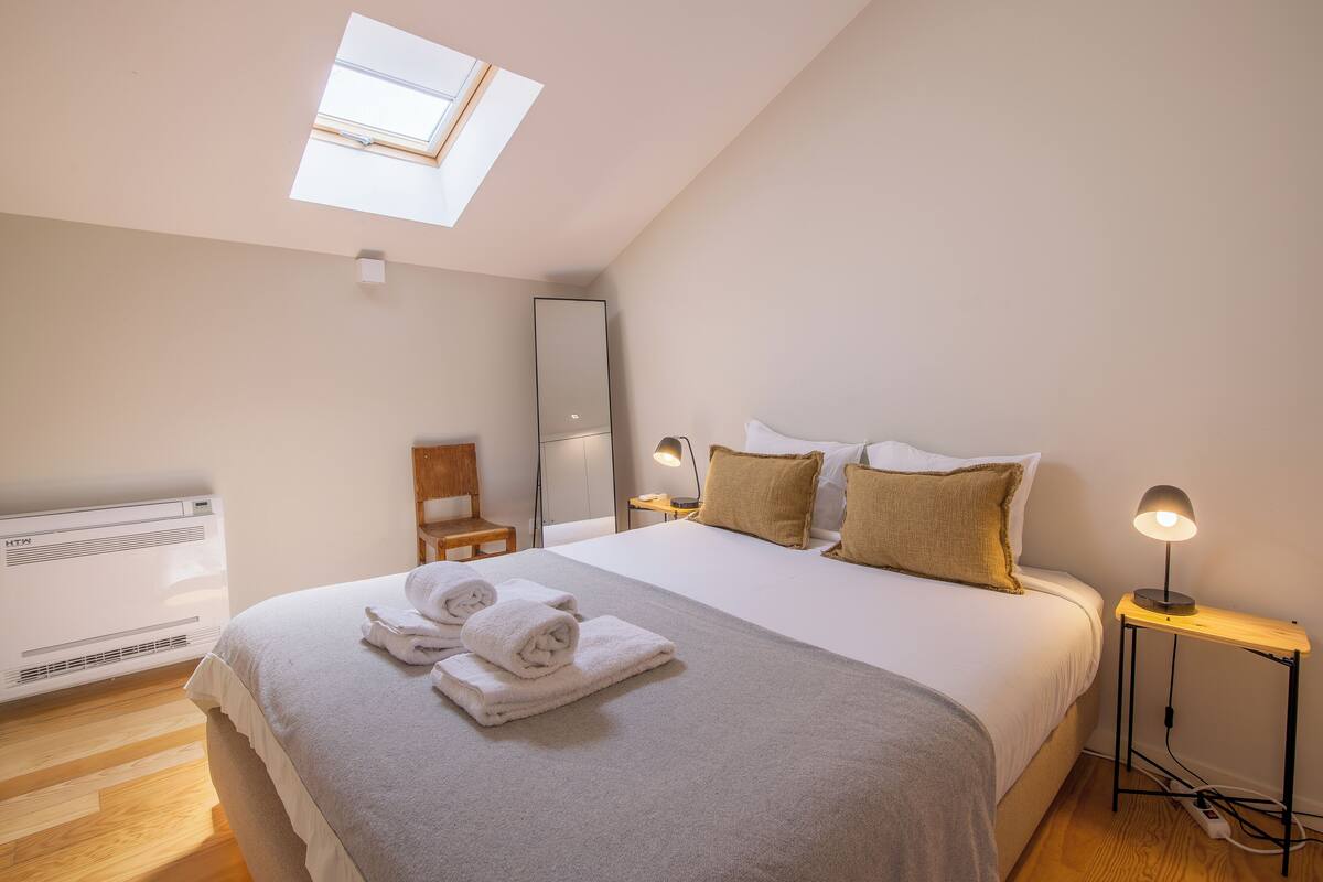 A bedroom with a skylight, a bed with white linens, a bedside table with a lamp, and a chair.