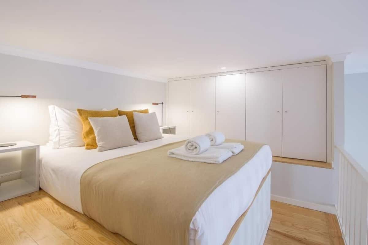 A bedroom with a bed, white walls, a wooden floor, a wardrobe, and a nightstand.
