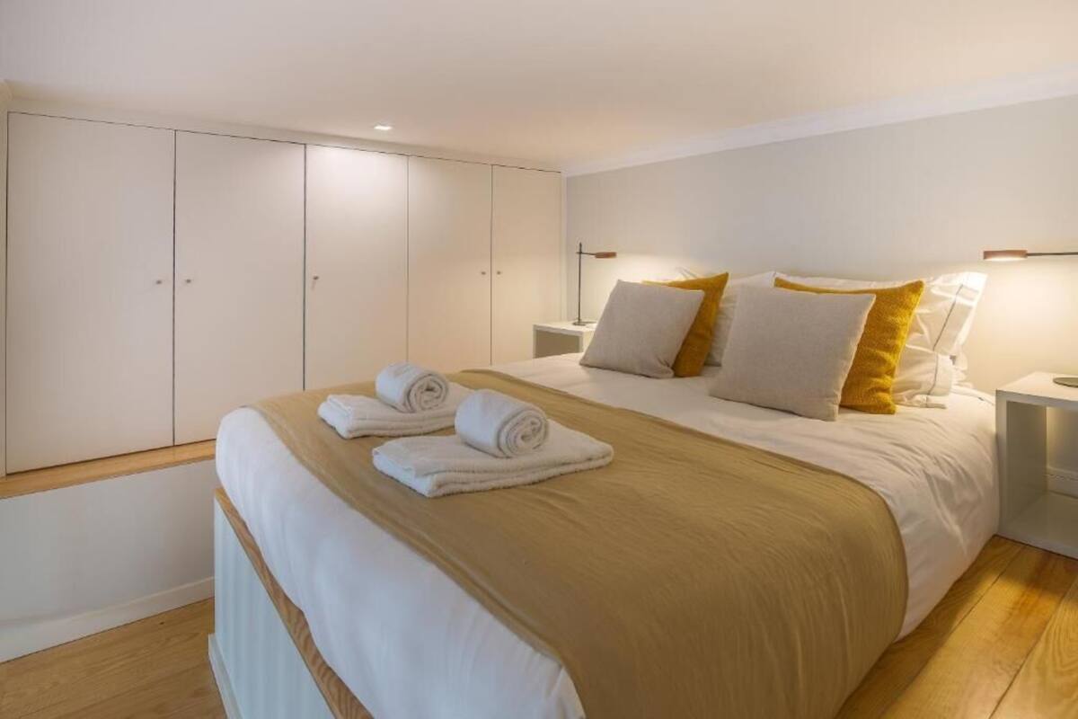A bedroom with a large bed, white and yellow pillows, a beige blanket, and neatly folded towels. There is a wardrobe with sliding doors and a bedside table with a lamp.
