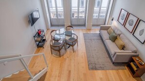 A modern living room with a sofa, a glass table with chairs, a TV mounted on the wall, and large windows with curtains. - ALMADA 151 (Porto)