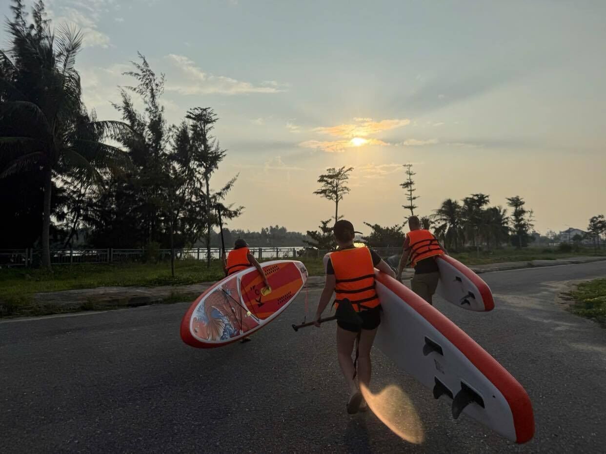 Dos personas caminando por una carretera con tablas de surf, usando chalecos salvavidas, y un atardecer de fondo.