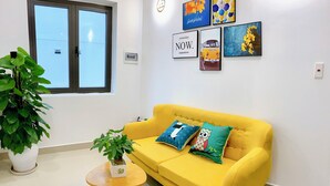 A yellow sofa with two pillows, a small round wooden table with a plant, and a large window with blinds. - EasyLife Villa Hoi An (Da Nang)