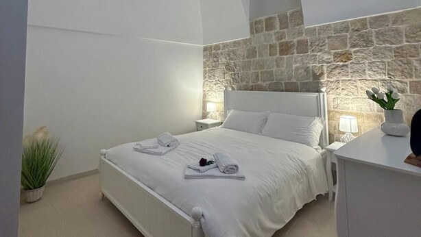 Un dormitorio con pared de piedra, cama blanca y mesita de noche con lámpara.