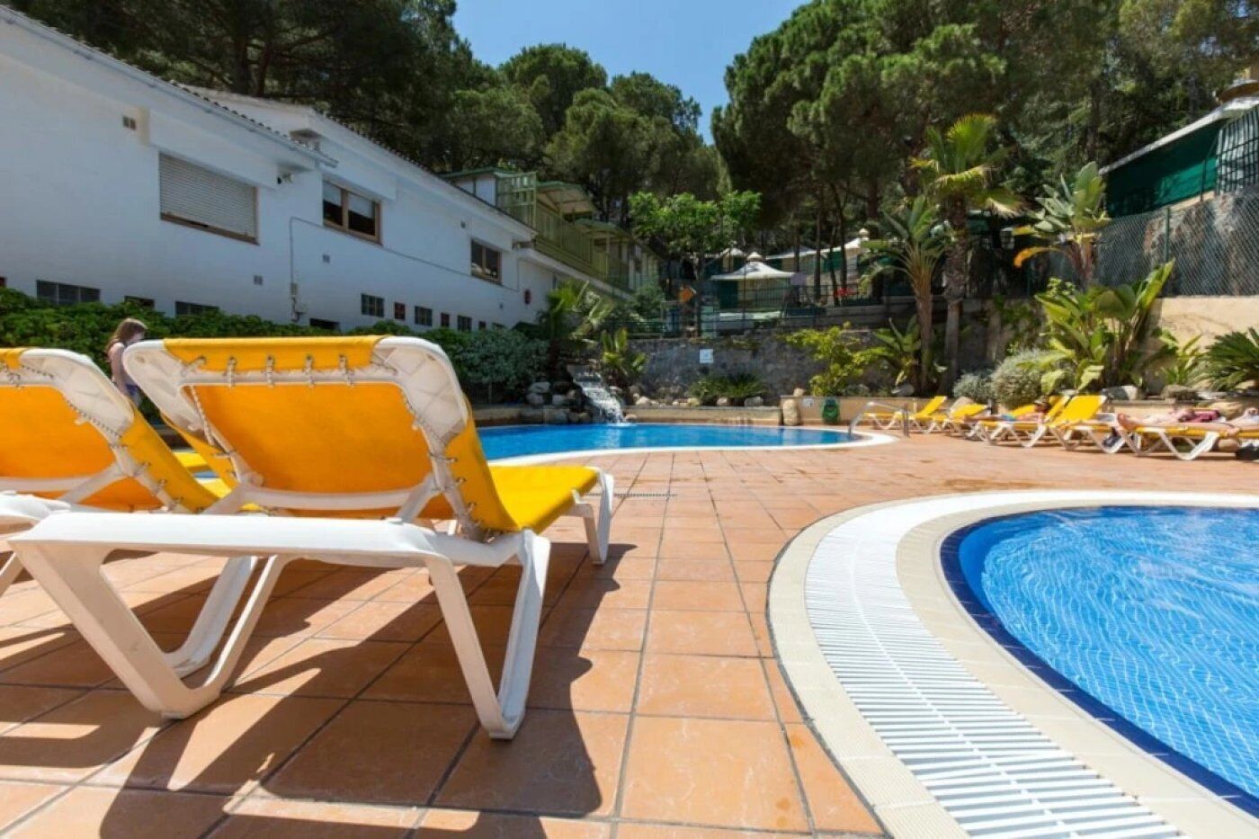 A pool area with yellow and white lounge chairs, a clear blue pool, and a white building in the background.