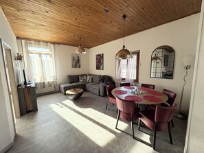 Ein modernes Wohnzimmer mit Holzboden, einem grauen Ecksofa, einem runden Esstisch mit roten Platzsets und einem großen Spiegel an der Wand.