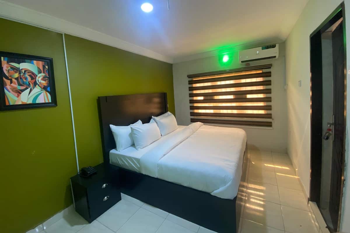 A modern hotel room with a large bed, a bedside table, a wall-mounted air conditioner, and a painting on the wall.