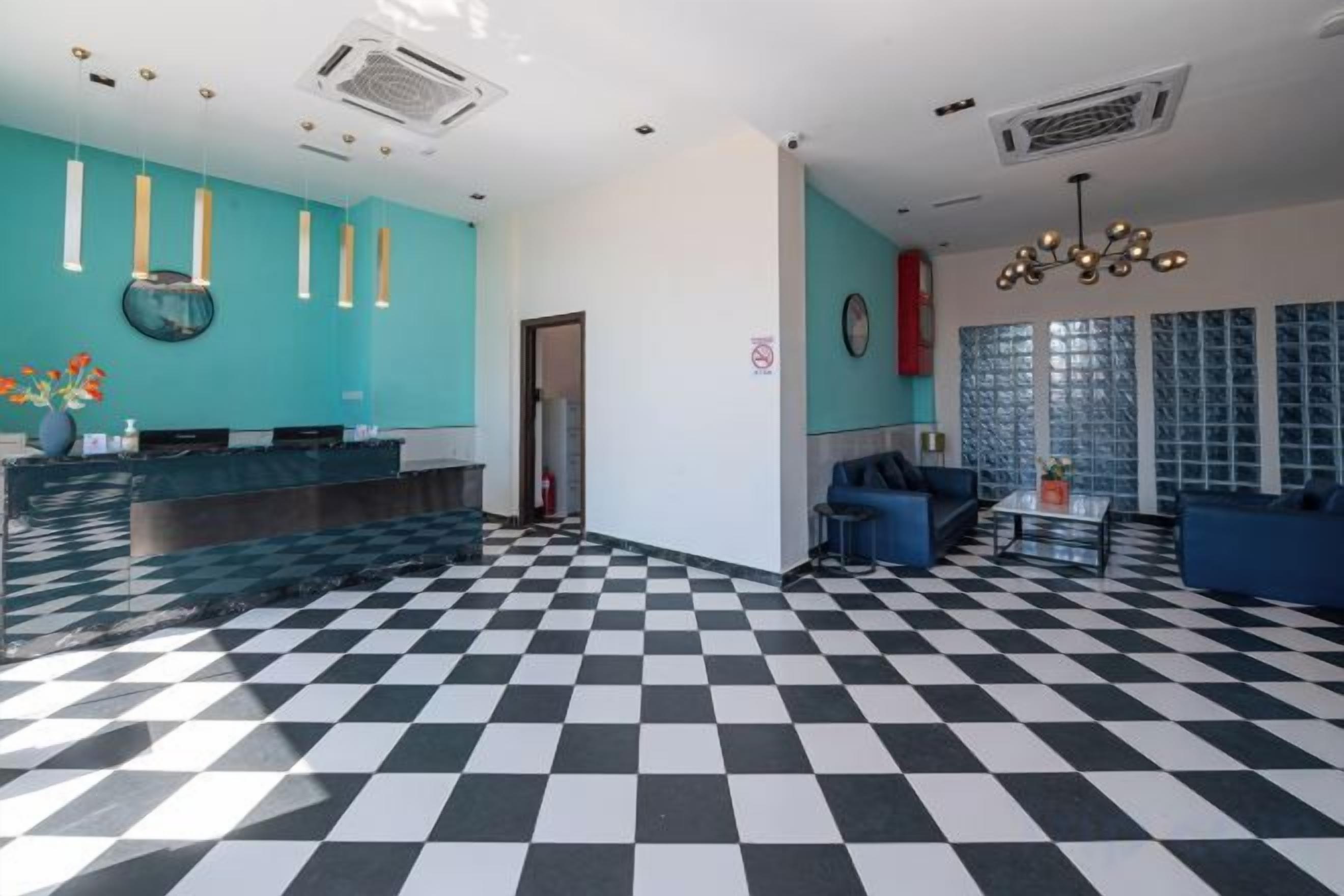 Un hall d’hôtel avec un sol à carreaux, un comptoir d’accueil, un espace salon et une grande fenêtre avec des stores.