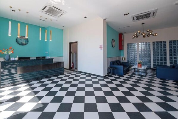 Una hall d'albergo con pavimento a scacchi, una reception, un'area salotto e un'ampia finestra con persiane.
