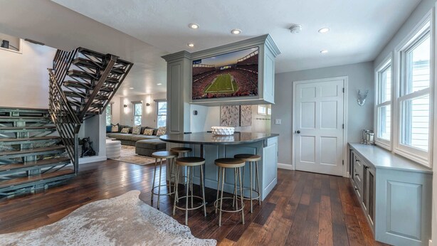 A spacious living area with a modern staircase, a bar with stools, and a large TV screen.