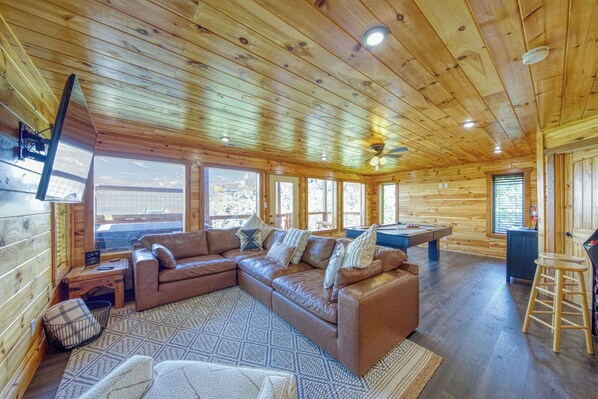A cozy cabin interior with a sectional sofa, a pool table, and large windows offering views. - 5-Star Mountain Views • Hot Tub • Blackstone • Game Room • Near Dollywood (Sevierville)