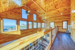 A wooden cabin interior with multiple windows offering views of a distant town and landscape. - 5-Star Mountain Views • Hot Tub • Blackstone • Game Room • Near Dollywood (Sevierville)