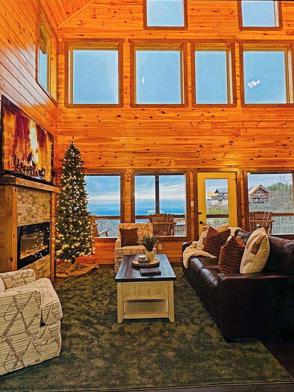 A cozy cabin interior with a fireplace, a Christmas tree, a leather sofa, and large windows offering a view of the outdoors.