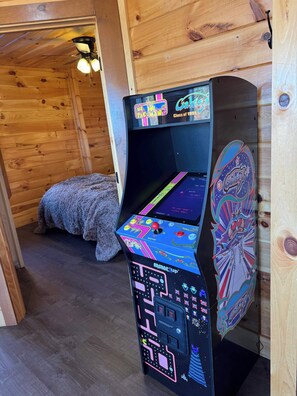A retro arcade machine with a colorful design, located in a wooden room with a cozy bed and a ceiling fan. - 5-Star Mountain Views • Hot Tub • Blackstone • Game Room • Near Dollywood (Sevierville)