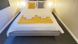 Un lit bien fait, recouvert d’une couette blanche, avec deux oreillers et un plaid jaune. On y trouve deux lampes de chevet et une œuvre d’art encadrée au-dessus du lit.