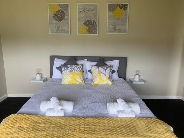 Un lit bien fait, recouvert d’une couvre-lit grise et jaune, avec deux oreillers ornés de motifs de feuilles et un plaid jaune. On y trouve deux tables de chevet avec de petites lampes.