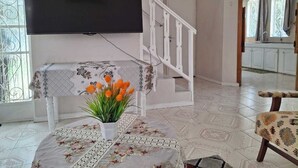 Un salon doté d’un téléviseur à écran plat fixé au mur, d’un escalier blanc, d’une table à manger recouverte d’une nappe à motifs floraux et d’une chaise agrémentée d’un coussin à motifs.