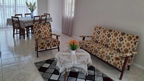 Un salon comprenant un canapé, deux fauteuils, une table à manger avec des chaises, un tapis à motifs et une petite table sur laquelle est posé un vase de fleurs.