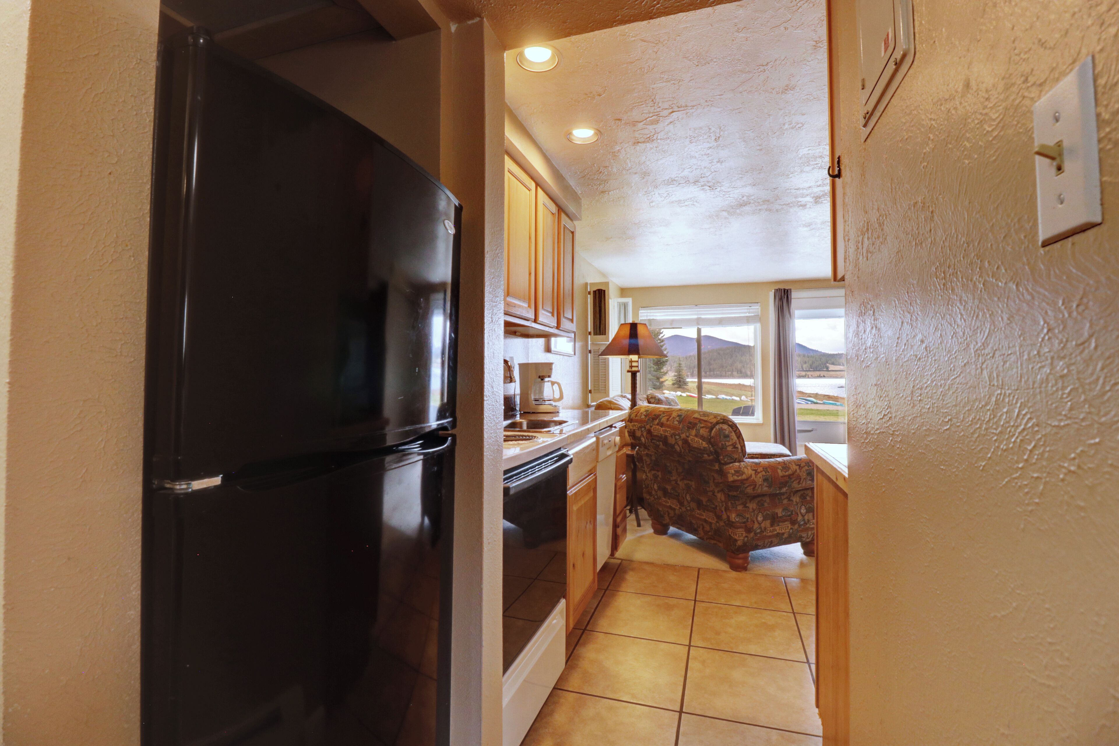 A compact kitchen with a black refrigerator, wooden cabinets, a patterned sofa, and a window offering a view of the outdoors.