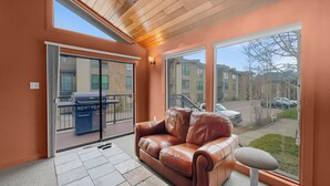 A room with a brown leather sofa, a glass door leading to an outdoor patio with a barbecue grill, and a view of a parking lot and buildings outside.