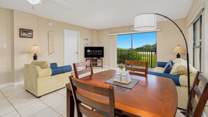 A spacious living room with a dining table, wooden chairs, a sofa, and a television. - Ormond Beach Oceanfront Accessible | Pool | Seabridge #101 (Ormond Beach)