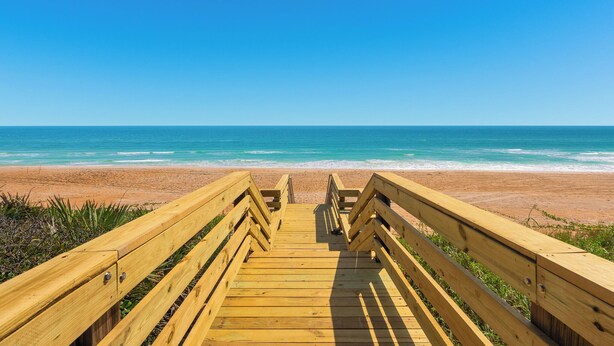 A wooden boardwalk leading to a beach with clear blue water and a sandy shore. - Ormond Beach Oceanfront Accessible | Pool | Seabridge #101 (Ormond Beach)