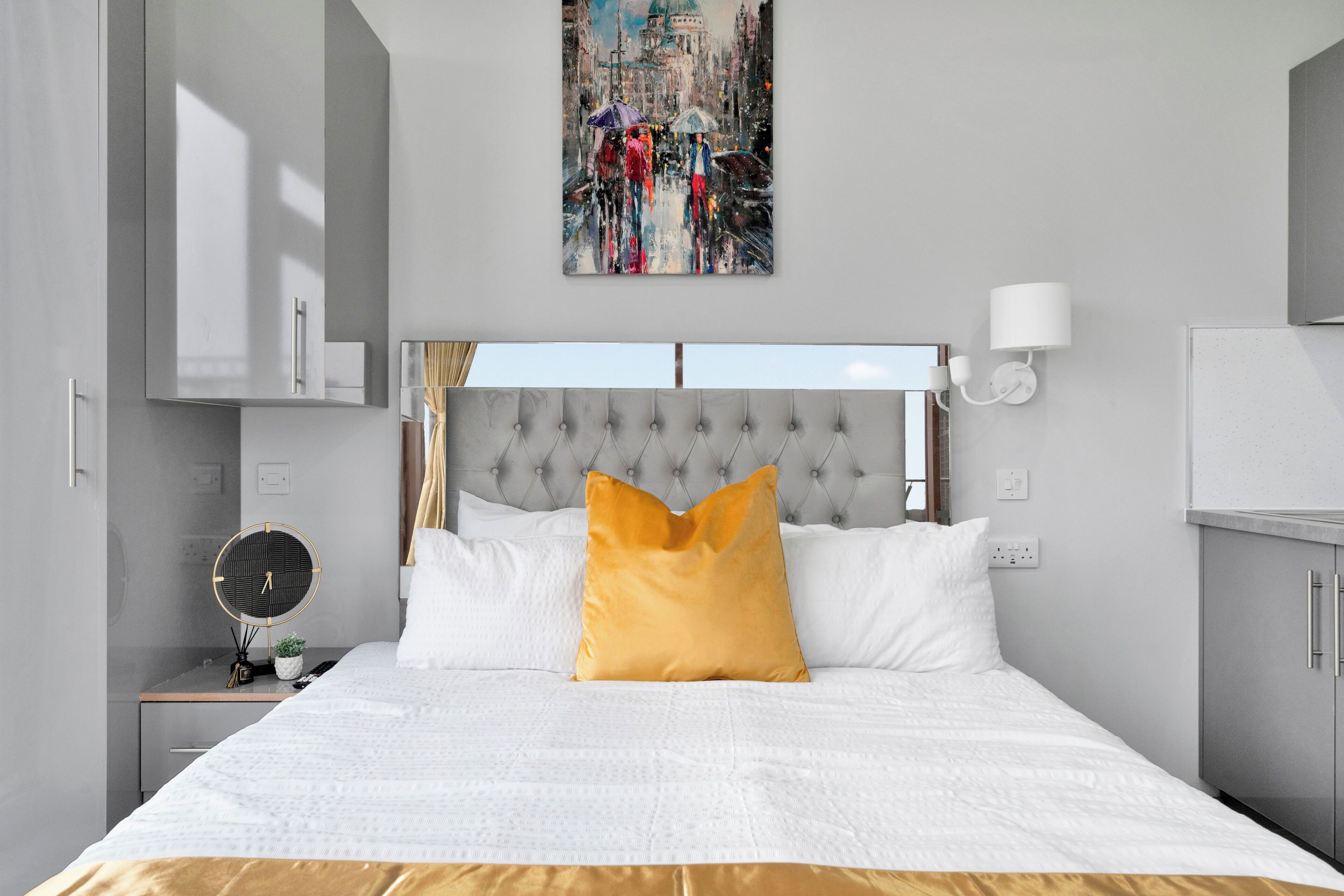 A modern bedroom with a tufted headboard, a bed with white and gold bedding, a round mirror, and a cityscape painting above the bed.