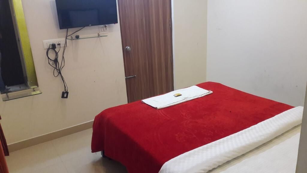A small room with a single bed covered in a red blanket, a white pillow, and a small table with a book. There is a wooden door and a wall-mounted TV.