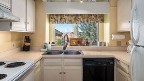 A kitchen with a stove, sink, and dishwasher, a window with a floral curtain, and a view of trees and buildings outside. - Bright, Spacious Ski Condo Close to the Resort! (Steamboat Springs)