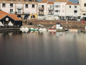 A waterfront with several boats docked, a small white building with a red roof, and multi-story buildings with balconies in the background.
