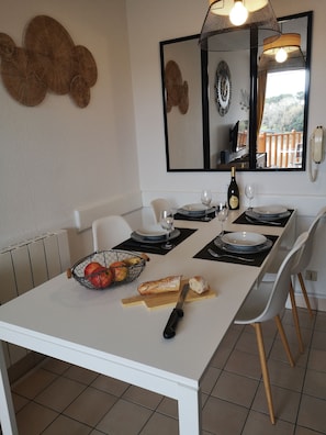 A dining area with a white table set for two, a fruit basket, a baguette, and a bottle of wine.