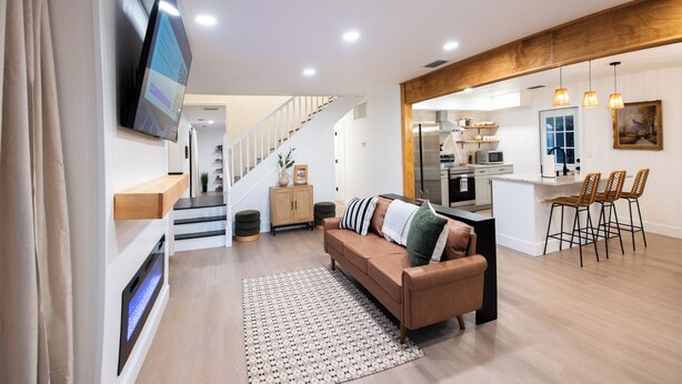 A modern living room with a sofa, a flat-screen TV, and a staircase leading to a kitchen area. - Vero Beach Gem: Pool, Game Room, King Beds, minutes to the beaches... (Vero Beach)