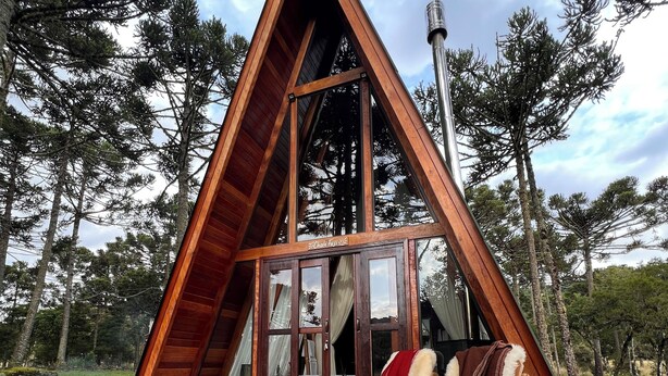 Una cabaña de madera con techo a dos aguas, un amplio ventanal, terraza de madera y zona de estar exterior.