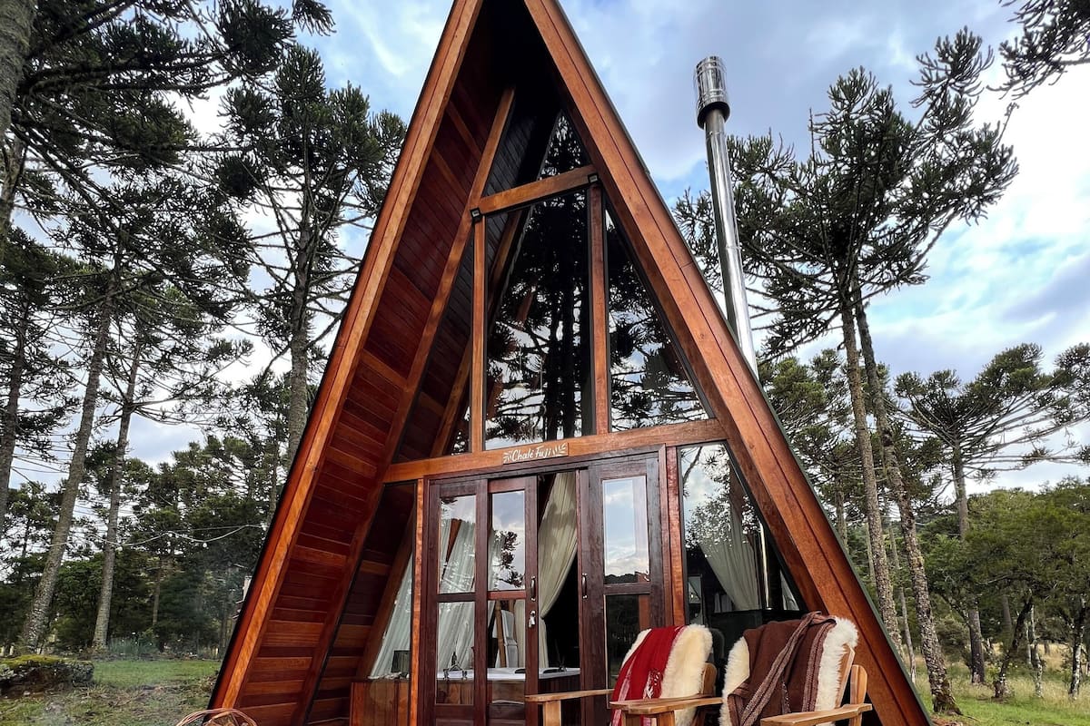 Une cabane en bois de forme triangulaire, dotée d’une grande porte vitrée, d’une terrasse en bois et d’un espace extérieur aménagé pour s’asseoir.