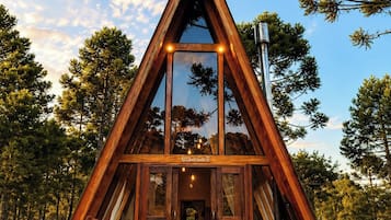 Una cabaña de madera triangular con un frente de vidrio, una hamaca, una mesa con una bebida y una fogata.