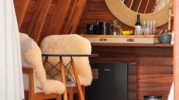 A cozy, well-lit room with a wooden ceiling, a plush chair, a small table with a bottle and glasses, a sink, and a trash bin.
