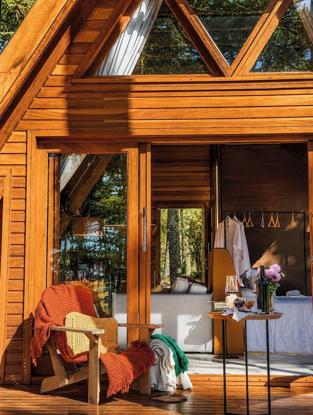 A wooden cabin with a glass door leading to an interior space. Inside, there is a seating area with a table, a lamp, and a bottle of flowers.