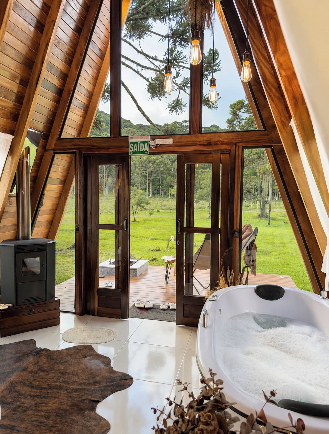 Interior com teto inclinado de madeira, hidromassagem e vista para uma paisagem verde através de grandes janelas.