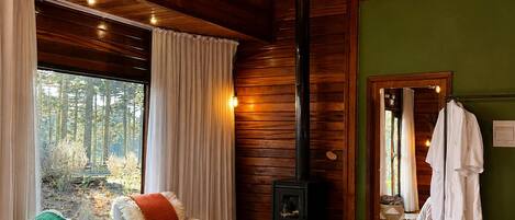 A cozy cabin interior with a bed, a sofa, a fireplace, and a large window overlooking a forest.