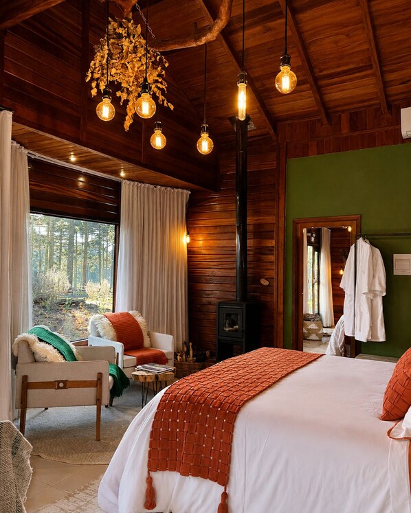 Interior acogedor de una cabaña con una cama, un sofá, una chimenea y un ventanal con vistas a un bosque.