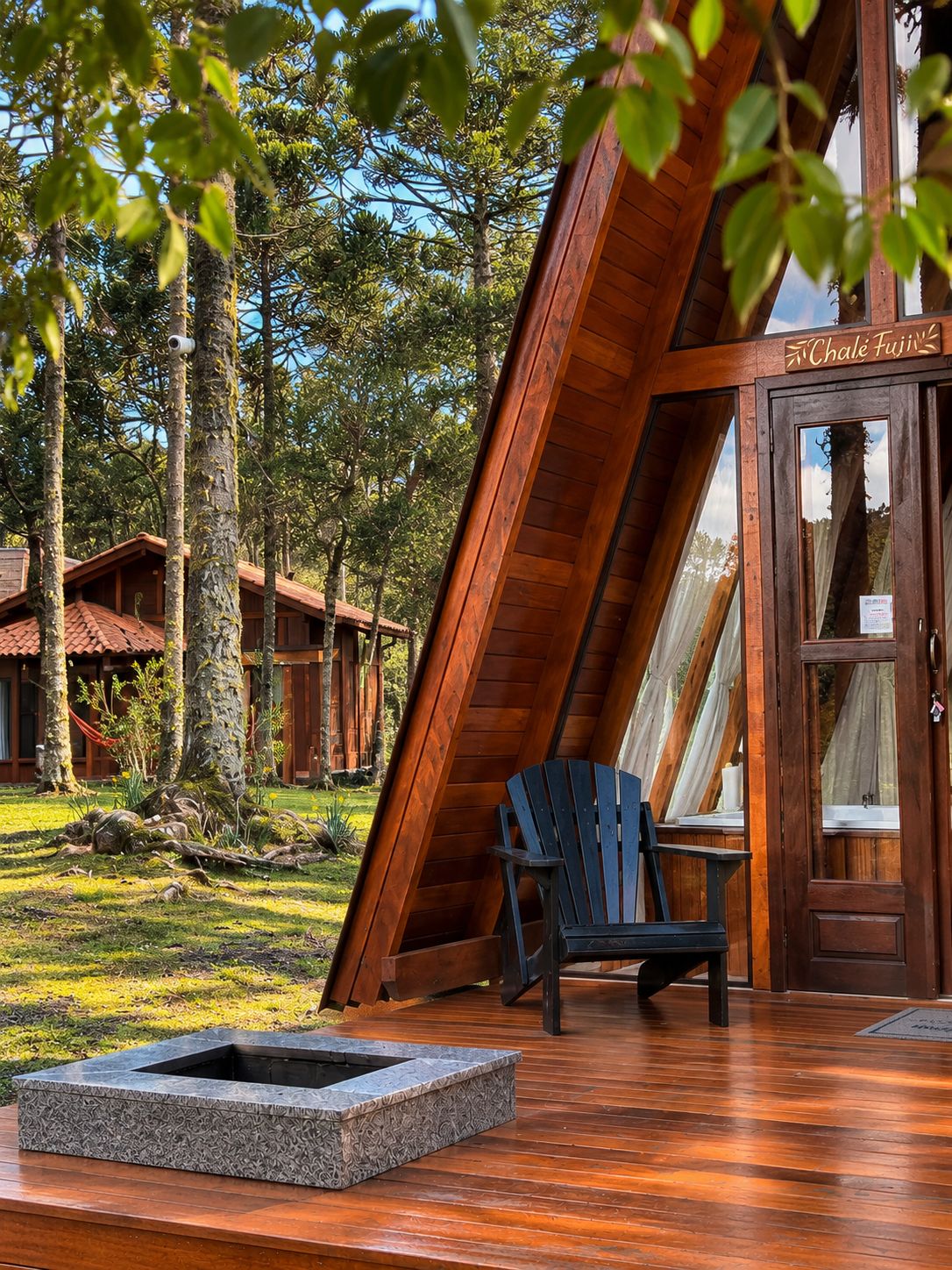 Una cabaña de madera con techo inclinado, una silla Adirondack negra y un brasero en una terraza de madera.