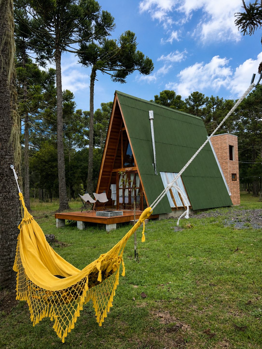 Uma pequena cabana de madeira triangular com telhado verde, cercada por árvores e uma rede.