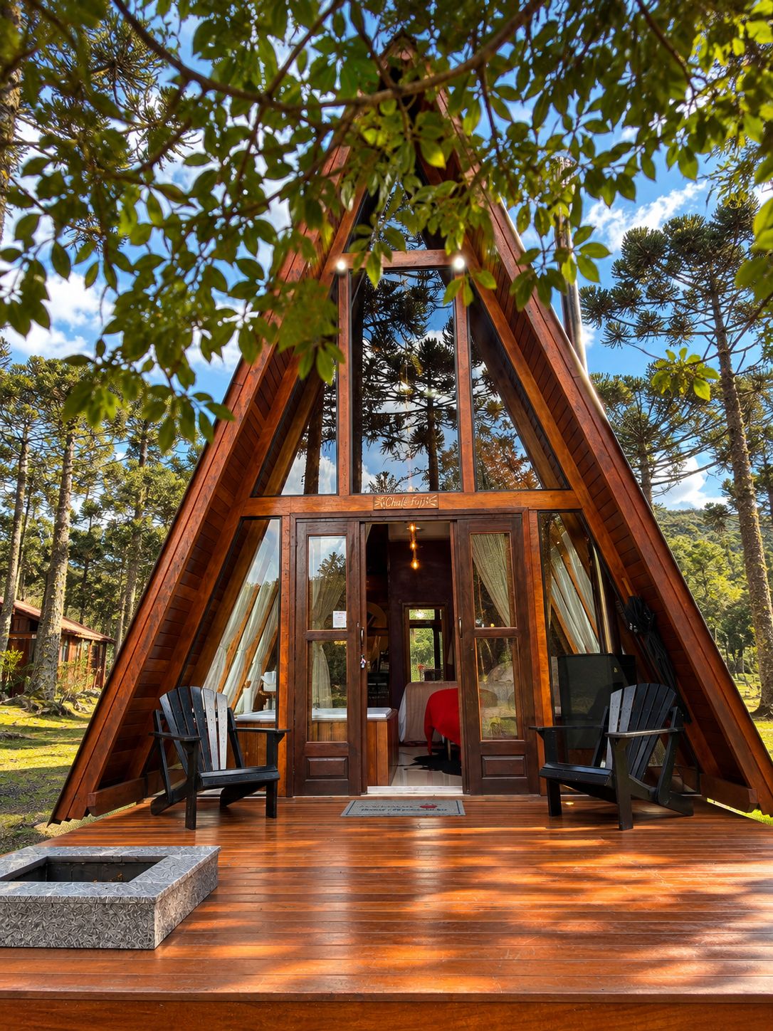 Uma cabana de madeira com telhado triangular, interior visível com cama e área de estar, e um deck com lareira.