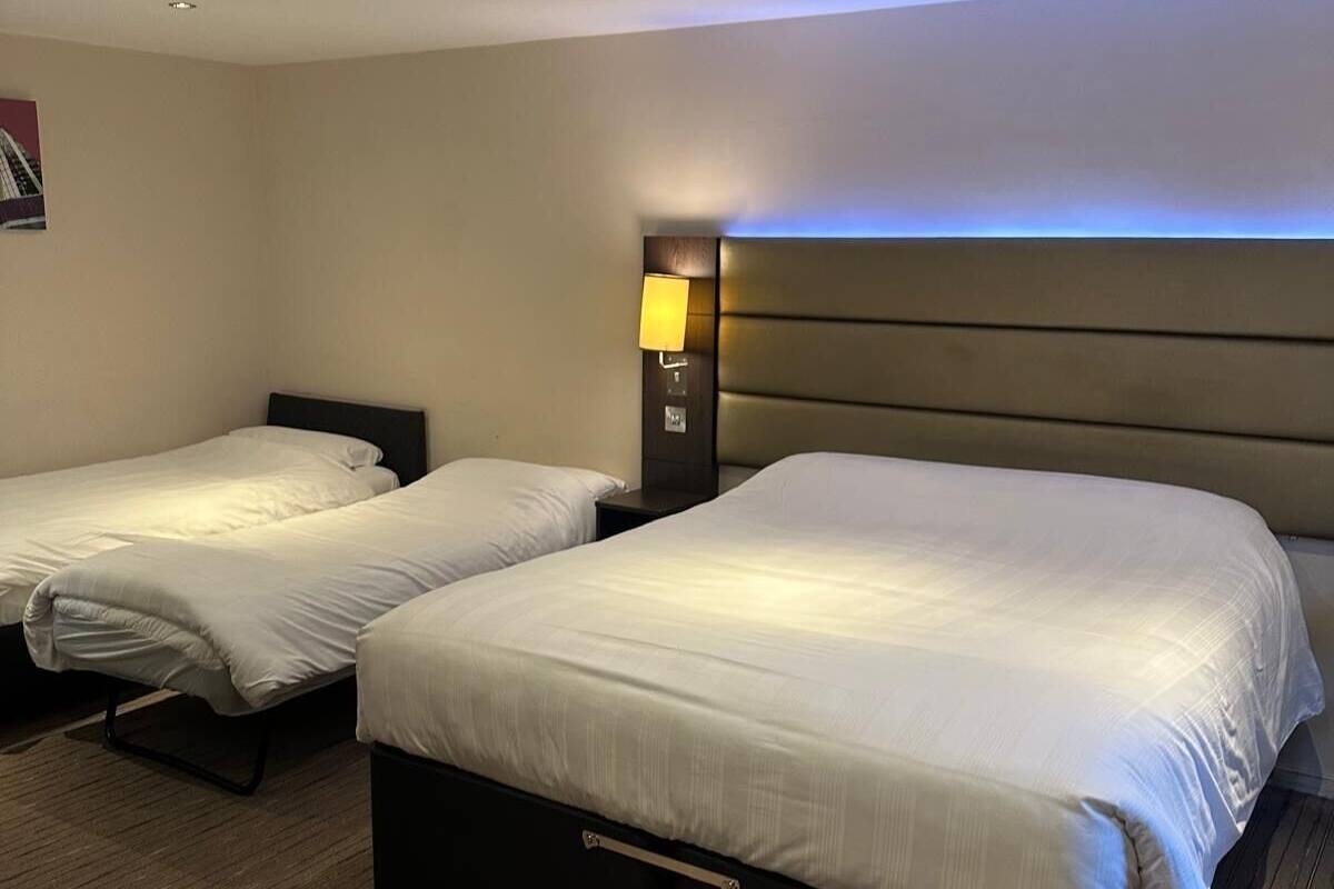 Quarto de hotel com duas camas de solteiro, uma mesinha de cabeceira com luminária e cabeceira com iluminação embutida.