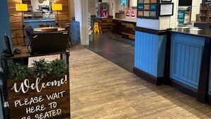 A restaurant interior with a wooden reception desk, a seating area, and a counter with menu boards. - Premier Roomz Two Mills (Ellesmere Port)