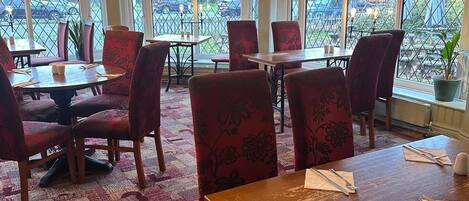 A restaurant with a high ceiling, patterned carpet, and red upholstered chairs. Tables are set with cutlery and napkins. Large windows offer views of the outside.